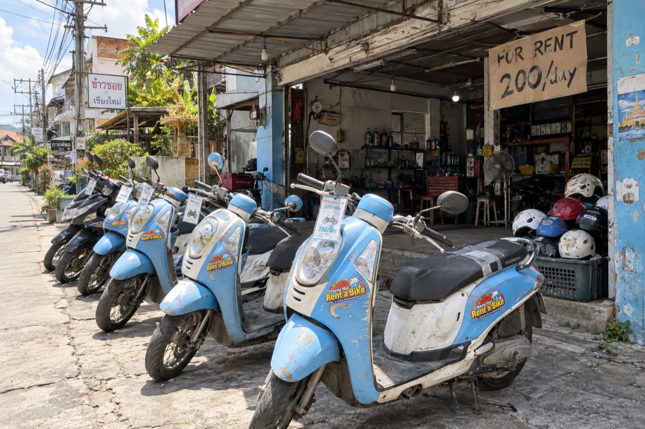 Typical scooter rental shop in Thailand: a row of aging baby blue 110cc scooters with scratched panels and big rental stickers, worn helmets piled in a crate, a handwritten 200 baht a day sign
